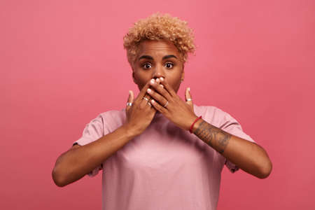 Scarred young dark skinned woman with frightened expression closes her mouth with hands. Female stands on pink background and feels fear and fright her eyes and gaze are full of fear and horror.の写真素材