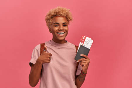 African american young female tourist holding tickets and showing thumbs up while standing on pink background. Passenger traveling abroad to travel on weekends getaway. People and vacation concept.の写真素材
