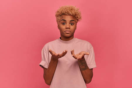 Portrait of confused dark skinned young woman with short blonde haircut spreading her arms and shrugging on pink background woman has doubts about choice. Concept of human feelings.の写真素材