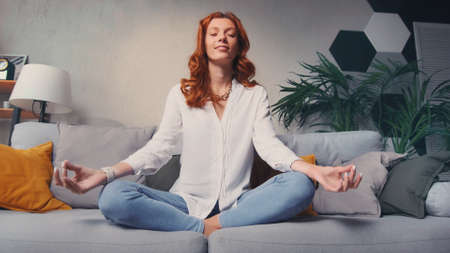 Woman sit cross legged on sofa in living room closed eyes do meditation practiceの写真素材