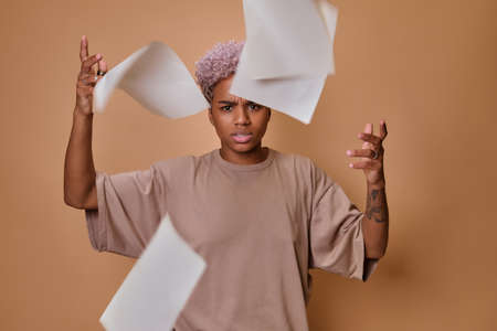 Angry sad African American woman throwing up documents on beige background.