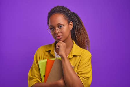 Young serious African American woman teacher touches chin and holds workbooks