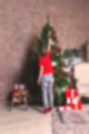 Blurred background with child in red t-shirt and socks standing on toes while decorating the Christmas tree. Holidays concept.の写真素材