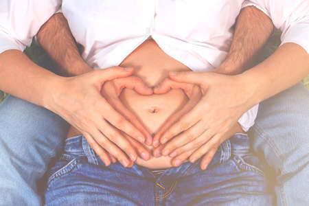 Young couple making heart shape on the pregnant belly. Minimalism lifestyle. Lovers in jeans and...