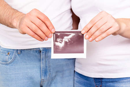 Dad and mom holding together in outstretched hands photo of their future baby. The baby's face is...