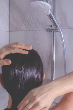 Photo of young woman from the back in shower. Unrecognizable woman washes her head. Jets of water flow from above.の写真素材