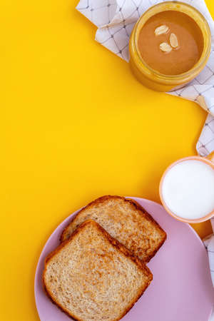 Top view of kid's breakfast. Appetizing toast on pink plate, peanut paste butter in jar and milk on vivid yellow backdrop and white napkin. Copy space.の写真素材
