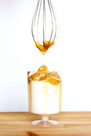 Dalgona coffee in glass on wooden table with cream on whisk. Creamy whipped trendy iced drink with...