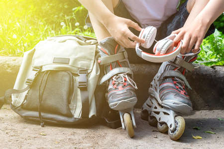 Unrecognizable caucasian boy with roller-skates, headphones and backpack sitting on sidelines to...