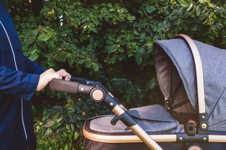 Mom in blue coat walking with her baby in park with grey stroller. Nature background. Side view. Close-up. Unrecognizable caucasian woman. Mom with prum. Baby carriage.の写真素材