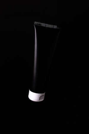Matte black cosmetic tube without label with white lid on black background. Monochrome color concept. Black tube object mock-up for packaging design. Poster design.の写真素材