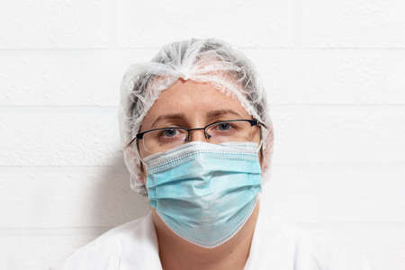 Close up of tired exhausted desperate caucasian female doctor in protective uniform and mask. After long shift at clinic doctor leaned against the wall to rest for a while.の写真素材