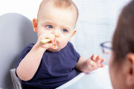 Cute caucasian baby boy is sitting in high chair and eating with plastic spoon. Mom is proud of her son, doesnt help him. Baby is eating on his own.の写真素材