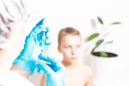 Hands in blue protective gloves. Doctor or nurse filling medicine from ampoule into syringe. Copy space. Light grey background. Close up. Focus on syringe. Doctor unrecognizable and blurred.の写真素材