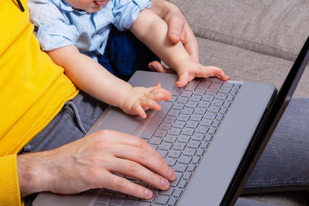 Small babys hands on laptop keyboard. Top view. Son helps daddy blogger work from home. Dad sitting...