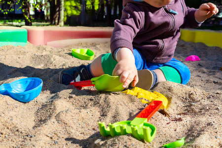 Unrecognizable little caucasian boy sitting in sandbox with toys on sunny summer day in big city....