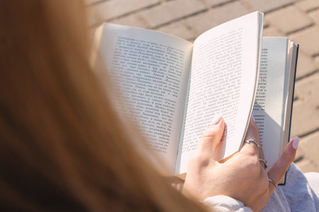 Unrecognizable caucasian woman in casual clothes sitting on park bench and reading real paper book....