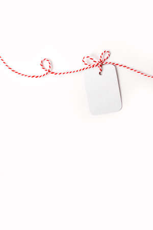 Small cardboard greeting card mock up on white-red ribbon on light background. Copy-space. Vertical format. Twists and bows on ribbon.の写真素材