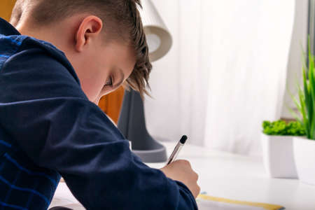 Attractive caucasian teenager with short hair in dark blue shirt doing his school homework sitting at white desk. Rear view. Studying, doing homework.の写真素材