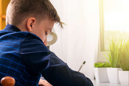 Attractive caucasian teenager with short hair in dark blue shirt doing his school homework sitting...