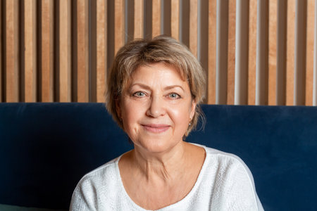 Beautiful elderly caucasian woman with short hair portrait. Woman in white t-shirt sitting on dark blue sofa.の写真素材