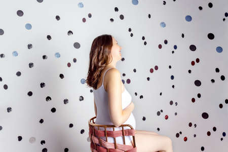 Pregnant caucasian woman with curly hair in white shirt sitting on chair and laughing. Polka dot wall background. Back rear view. Adorable pregnancy.の写真素材