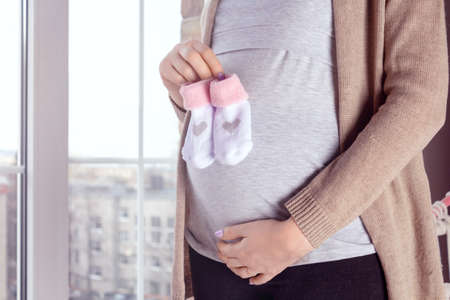Unrecognizable caucasian woman standing near the window and holding small pink socks for future newborn daughter in hands. Casual clothing. 28 week of pregnancy.の写真素材
