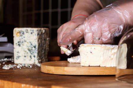 Cheese store. Unrecognizable saleswoman in apron slices crumbly blue mold Roquefort cheese with special knife on wooden board. Selective focus.の写真素材