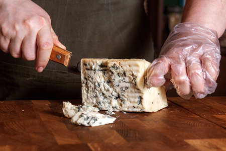 Cheese store. Unrecognizable saleswoman in apron slices crumbly blue mold Roquefort cheese with special knife on wooden board. Selective focus.の写真素材