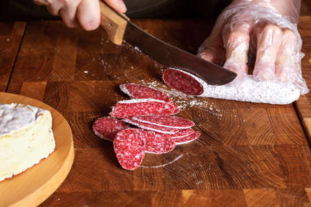 Saleswoman wearing disposable gloves slices salami sausage on wooden board. Small grocery store. Cheese shop. Hands with knife close-up.の写真素材