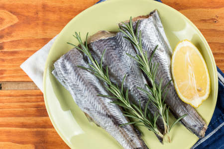 Hake fish with rosemary and lemon on the green plate above wooden board.