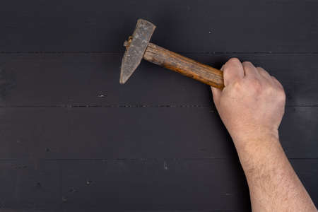 Old used hammer with wooden handle in the male hand above black wooden boards table. DIY concept with copy space above black wooden background.の写真素材