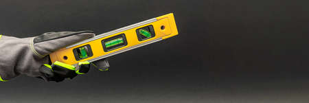 Hand in gloves holds a yellow building level on a black background. Postcard or poster for print, place for textの写真素材