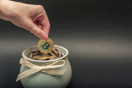 Investments, accumulation in cryptocurrency. Bitcoin in hand, female hand puts a gold bitcoin coin into a pot of coins. digital money, place for textの写真素材