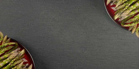Green asparagus on a black slate stone background. Top view, asparagus in a red plate. Culinary banner with place for text.の写真素材