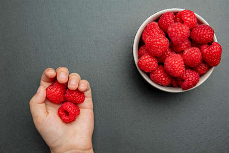 Raspberries in a saucer on a black background. The child takes raspberries from the plate and puts...