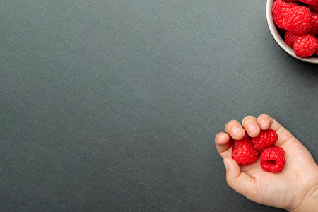 The child takes a raspberry and puts it in his hand. Berries on a black background in a plate, place to insert text, recipe or designの写真素材