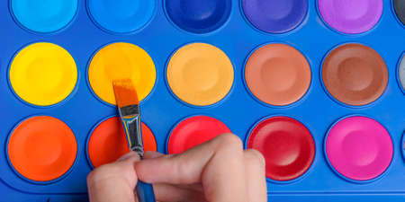 A set of watercolor paints and a brush for drawing close-up. Childs hand with a paintbrush. Selective focus.の写真素材