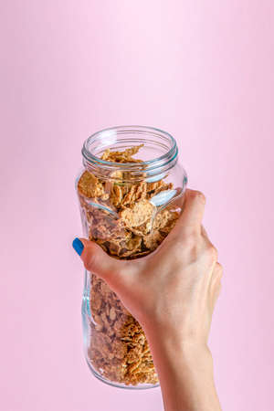 Muesli on a pink background. Pour dry muesli flakes by hand into a bowl. Pouring muesli from a glass jar into a black bowl when making cereals with muesli.の写真素材
