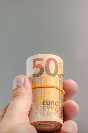 The hand holds a roll of 50 euro banknotes. Euro banknotes rolled up in a white mans hand on a gray background. The concept of financial assistance, real estate purchase, loan or insurance payment.の写真素材