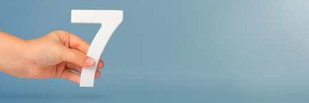 Number seven in hand. Hand holding white number 7 on blue background with copy space. Concept with number seven. Birthday 7 years, percent, seventh grade or day.の写真素材