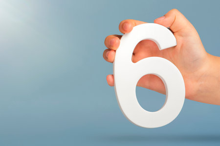 Number six in hand. A hand holds a white number 6 on a blue background with copy space. Concept with number six. 6 percent, birthday 6, sixth grade, in sixth gradeの写真素材