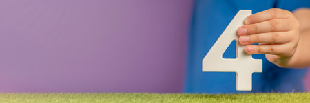 Number 4 in hand. Hand holding white number four over green grass and purple background, birthday concept, competition place, four percent per annum. Canada National Day, USA Independence Day, 4 July.の写真素材