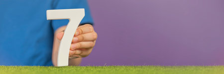 Number 7 in hand. A hand holds a white number seven on green grass and a purple background, seven percent per annum. World Health Day. April 7th. Holiday concept.の写真素材