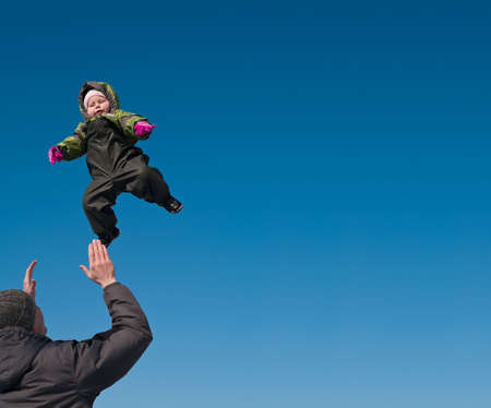 Father playing with little child. Man tosses up a daughter. On blue sky background. Winter clothes.の写真素材