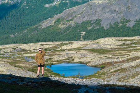 Man in mountain standing near lake and looking at camera from far awayの写真素材