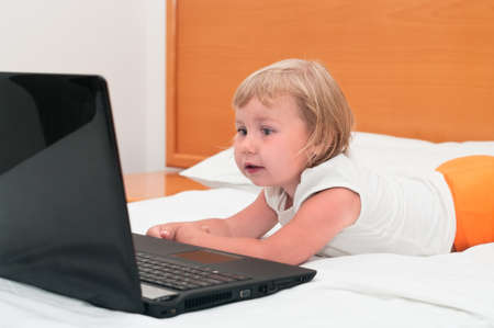 A small child is sitting with a laptop on the white and orange bedの写真素材