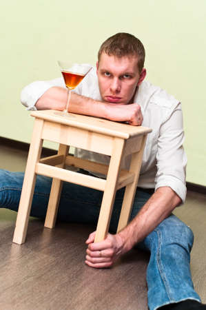 Drunken man sitting on floor with glass of alcohol and holding stoolの写真素材