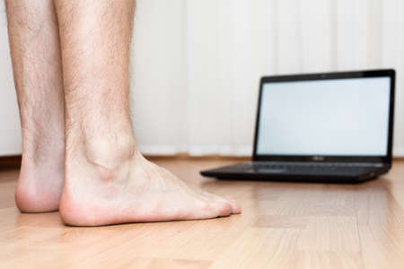 Opened laptop and male feet on the parquet flooring of the room. Laptop not in focus. Focus on legs.