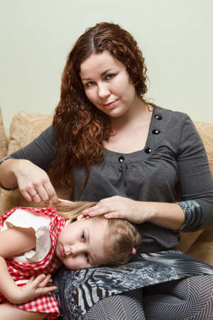 Mother and little daughter sitting on couch. Woman combing daughter hairの写真素材